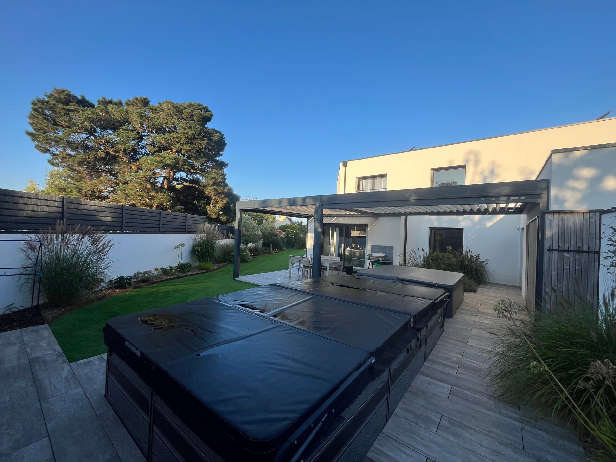 Terrasse moderne avec pergola bioclimatique, spa de nage et gazon synthétique aménagés par AJY Maçonnerie dans le jardin d’une maison contemporaine.