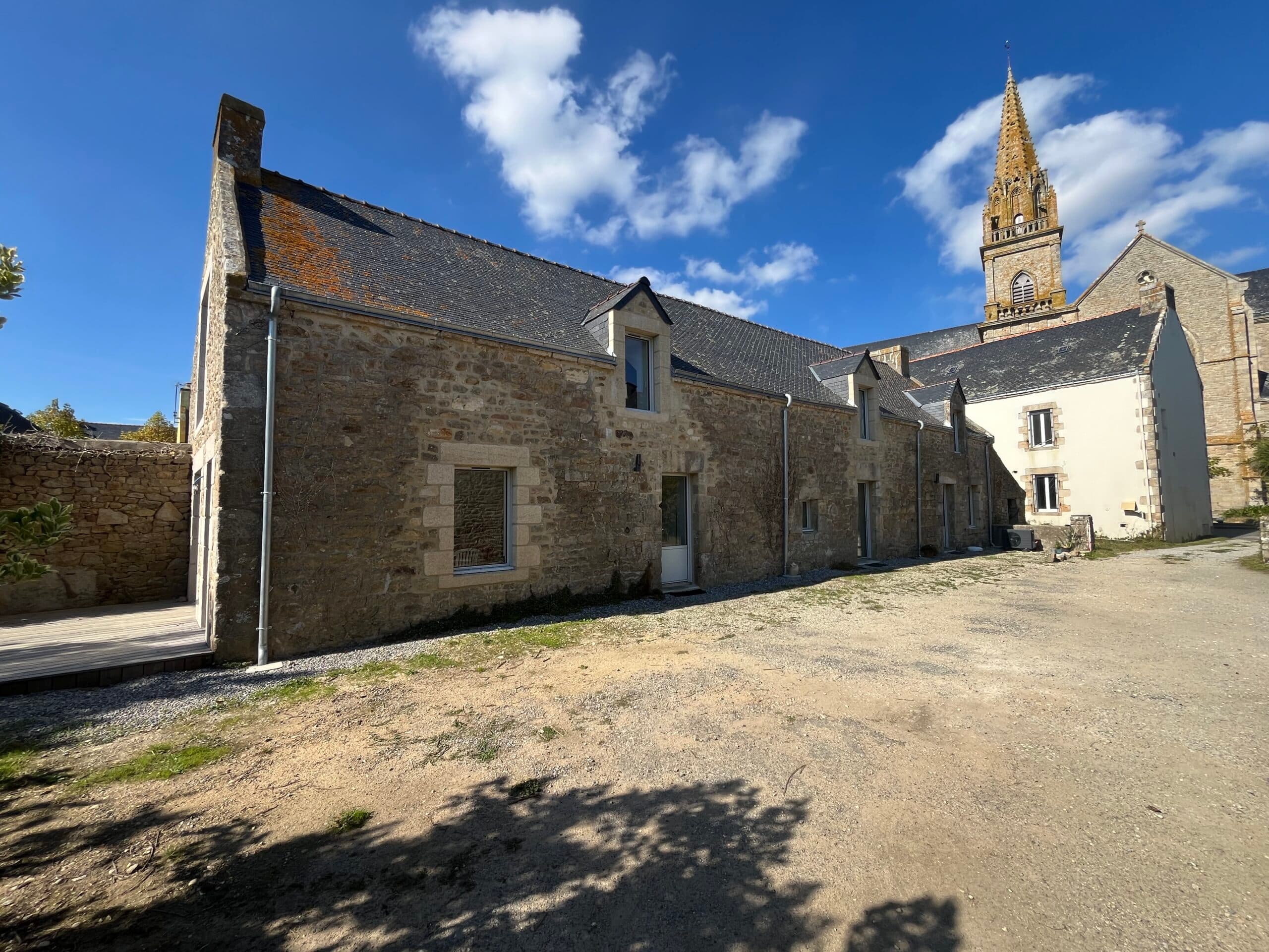 Maison bretonne en pierre rénovée par AJY Maçonnerie avec grande baie vitrée et terrasse bois créant un contraste moderne dans un jardin de village.