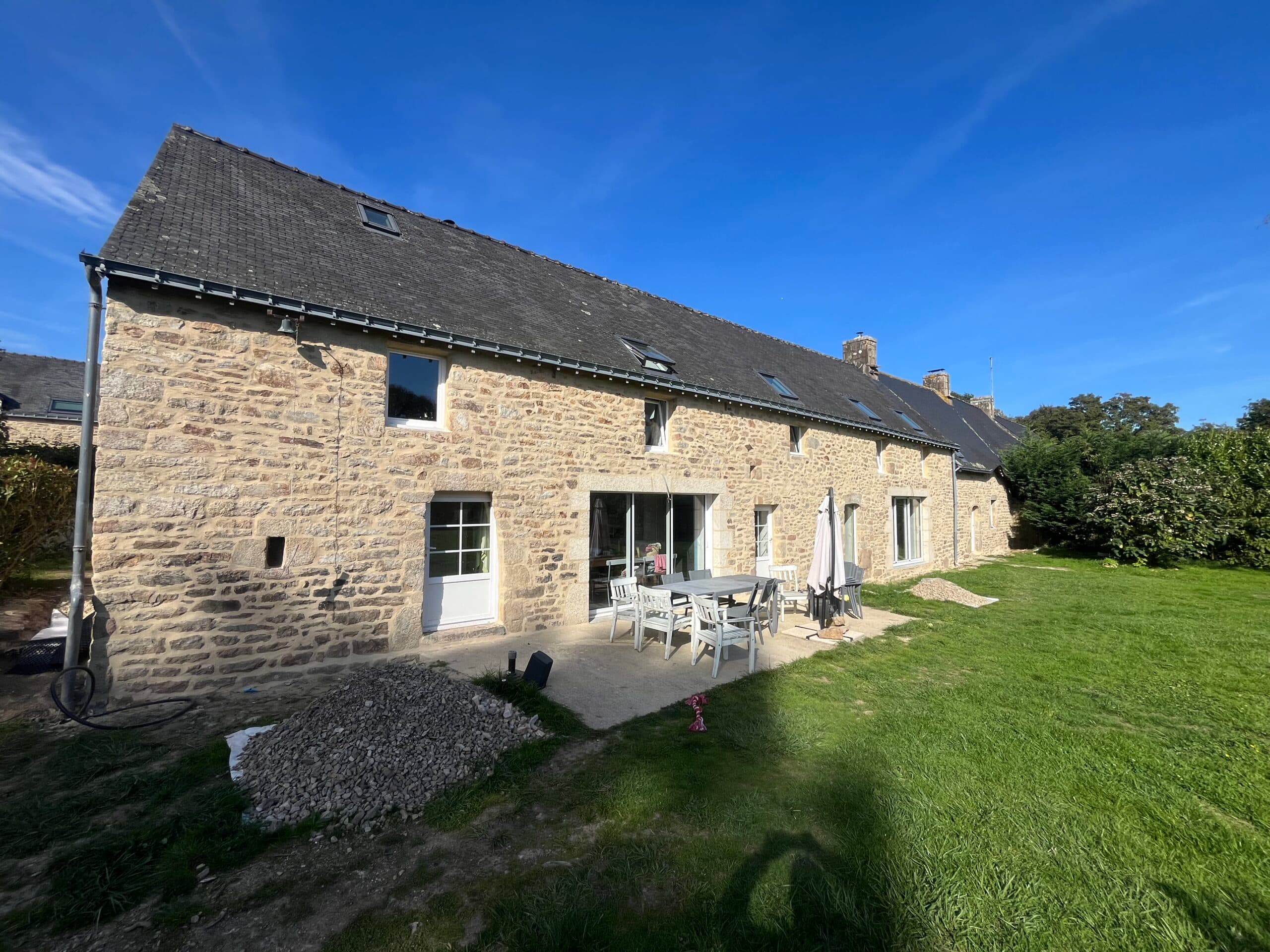 Vue large d’une longère en pierre rénovée par AJY Maçonnerie avec terrasse aménagée et grand jardin