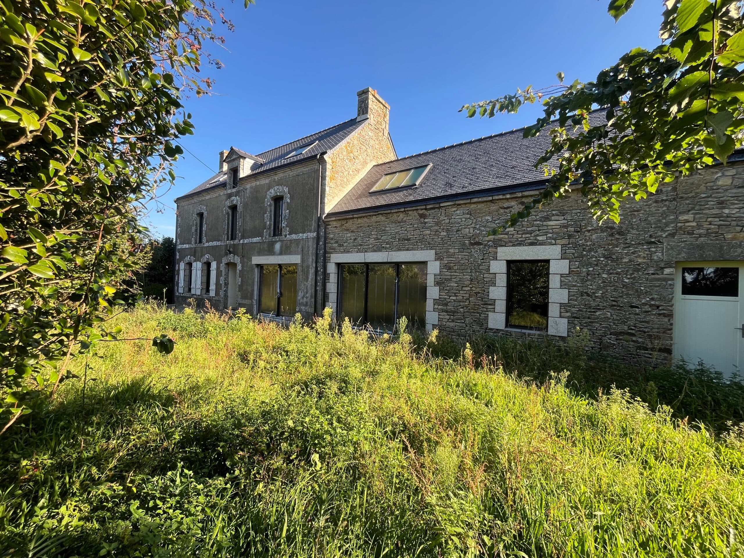 Façade sur rue d’une longère en pierre avec panneaux AJY Maçonnerie, illustrant un chantier de rénovation de maison bretonne avec garage intégré.
